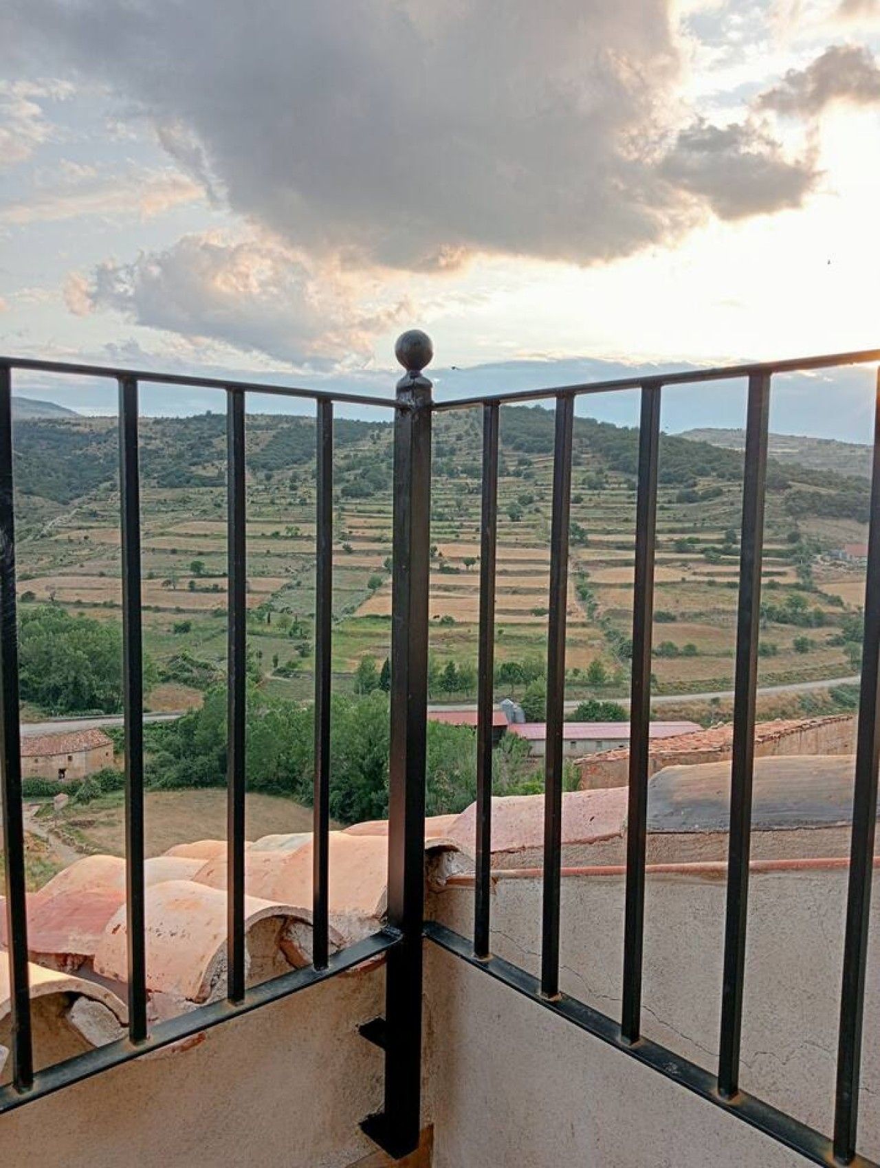 Een balkon met metalen hekwerk met uitzicht op een landelijk gebied met velden en verspreide gebouwen.