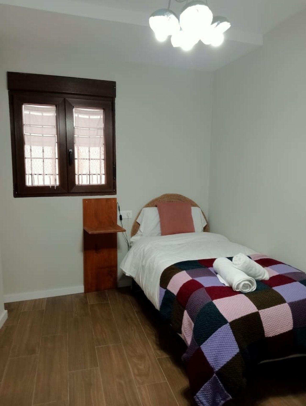 Una cama individual con una colcha de cuadros, una mesita de noche de madera, una ventana con persianas y una lámpara en el techo.