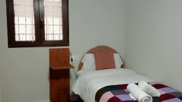 Una cama individual con una colcha de cuadros, una mesita de noche de madera, una ventana con persianas y una lámpara en el techo.