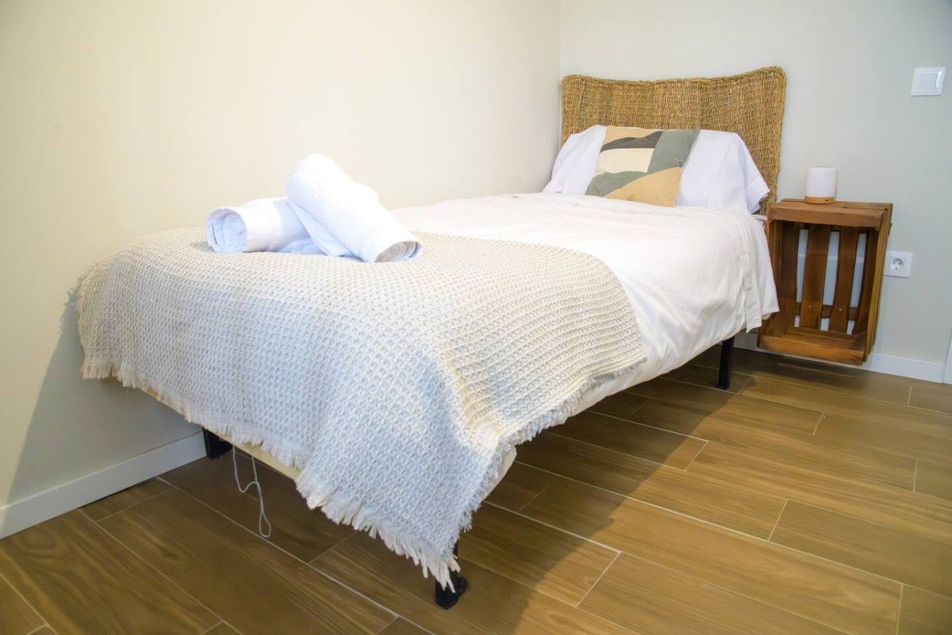 Una cama con una colcha blanca y una toalla enrollada, una mesita de noche de madera con una lámpara y un suelo de madera.