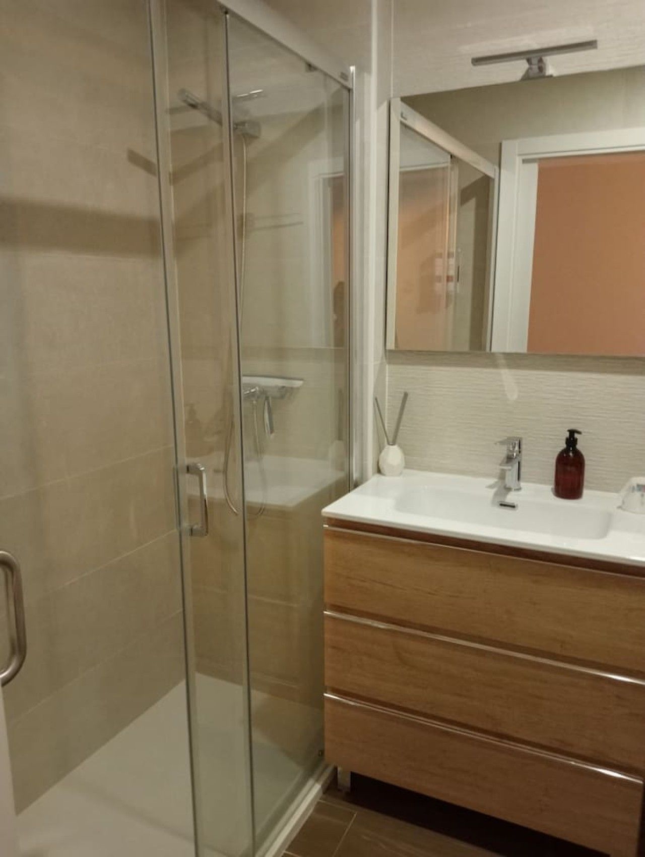 Een badkamer met een douchecabine van glas, een witte wastafel met zeepdispenser en een houten wastafelmeubel met lades.