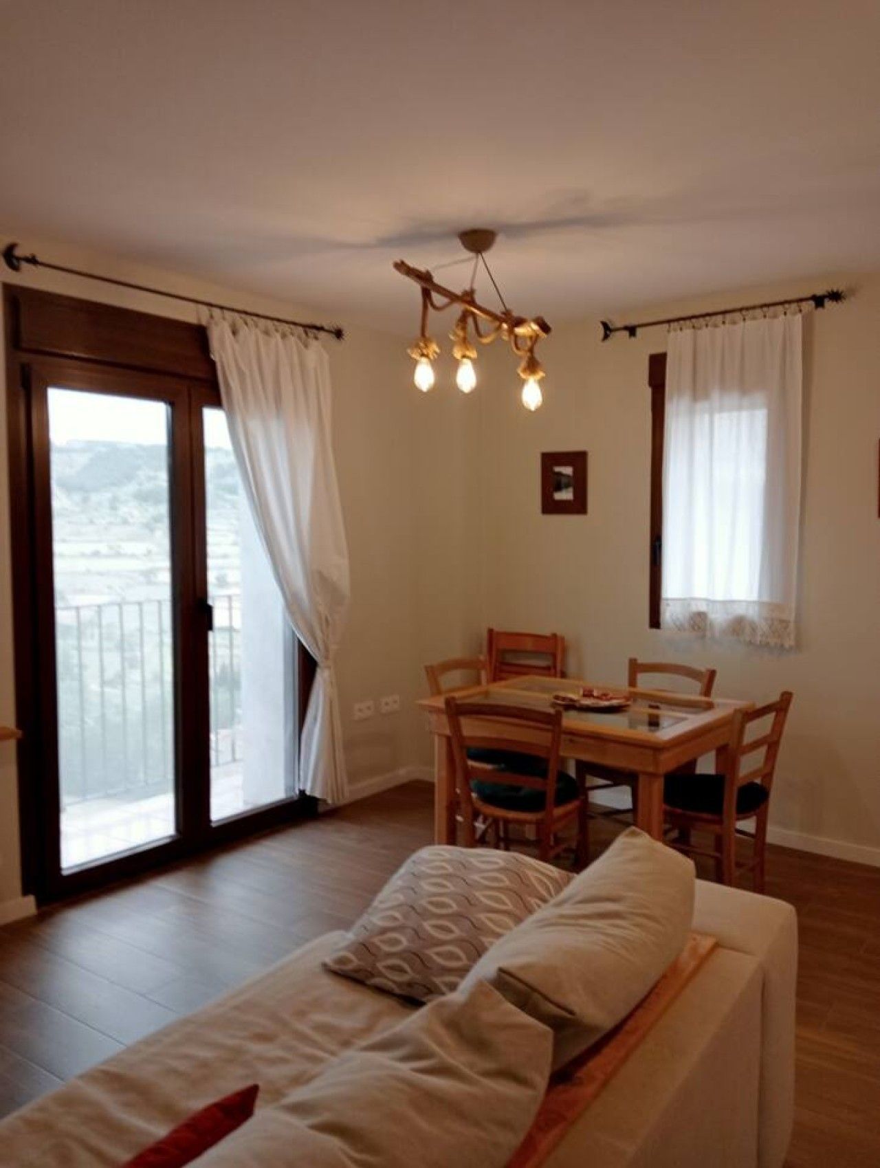 A room with a bed, a dining table with chairs, a ceiling light fixture, and a view through a door.