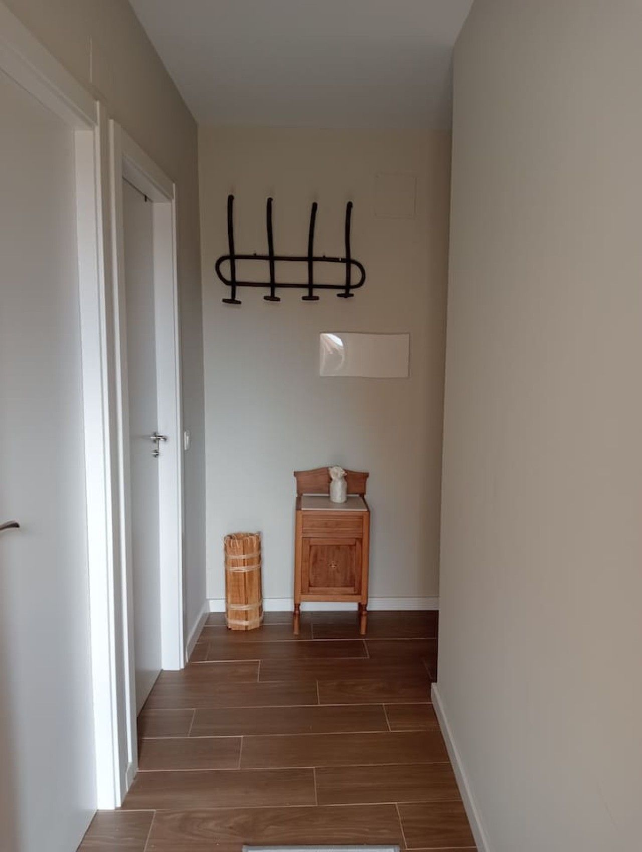 Un pasillo con suelo de madera, una puerta a la izquierda, una pequeña mesa de madera con un florero y un perchero en la pared.
