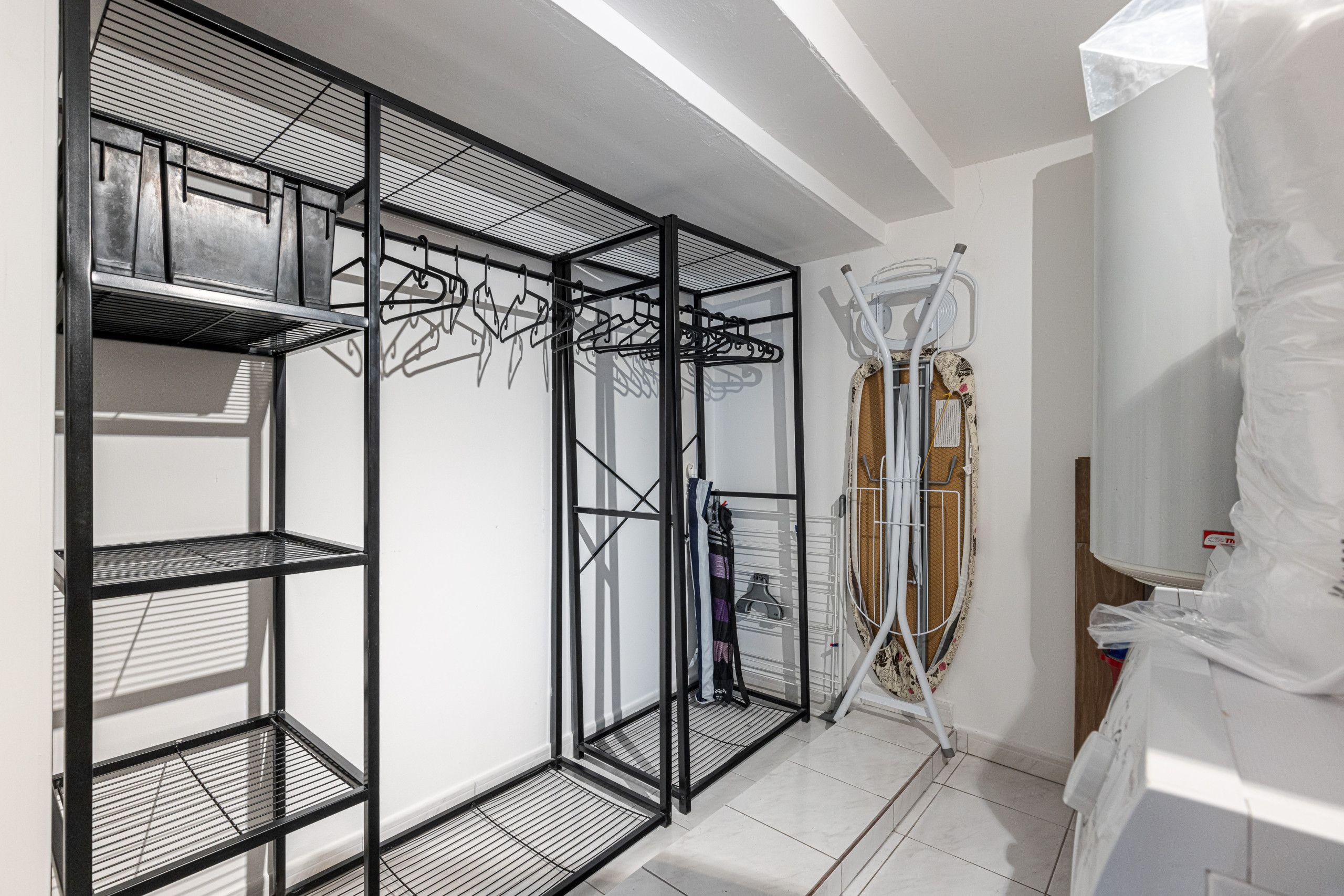 Ein modernes, mehrstufiges Metallregal mit Kleiderhaken, eine kleine Erhöhung zu einer höheren Ebene und ein weißer Vorhang, der einen weißen Heizkörper teilweise verdeckt.