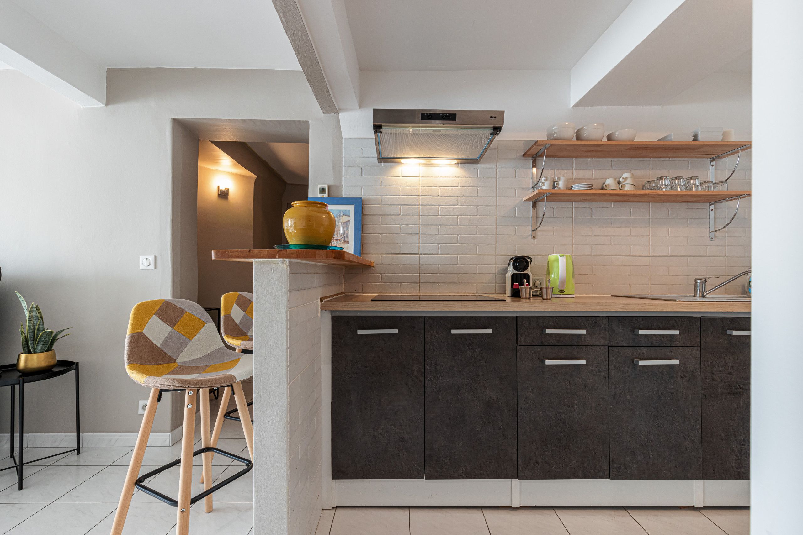 Une cuisine moderne avec des meubles de couleur sombre, un plan de travail en bois et un four encastré. On y trouve deux chaises à motifs géométriques, une plante en pot et une étagère contenant des ustensiles de cuisine.