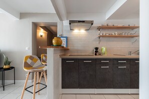 Een moderne keuken met donkere kastjes, een houten aanrechtblad en een ingebouwde oven. Er staan twee stoelen met een geometrisch patroon, een potplant en een plank met keukengerei.