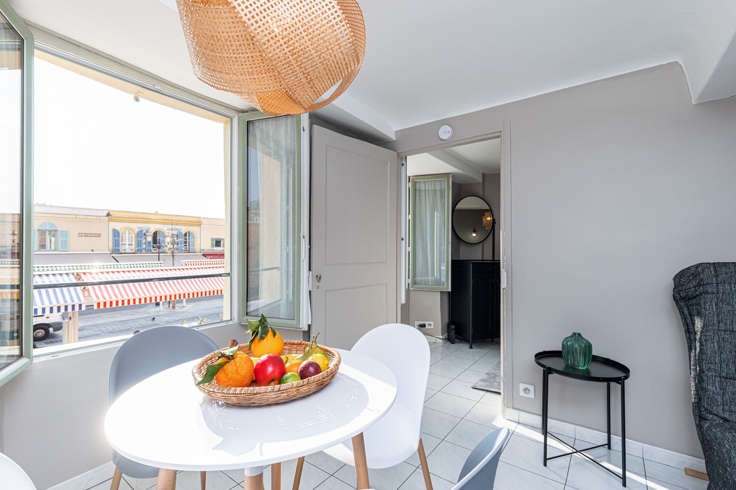 Un salon moderne avec un coin repas, une grande fenêtre donnant sur la rue et un panier de fruits posé sur la table.