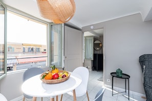 Een moderne woonkamer met een eethoek, een groot raam met uitzicht op de straat, en een mand vol fruit op de tafel.