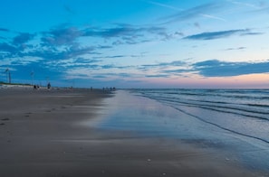 Uma praia com mar calmo, céu claro e turbinas eólicas ao longe.