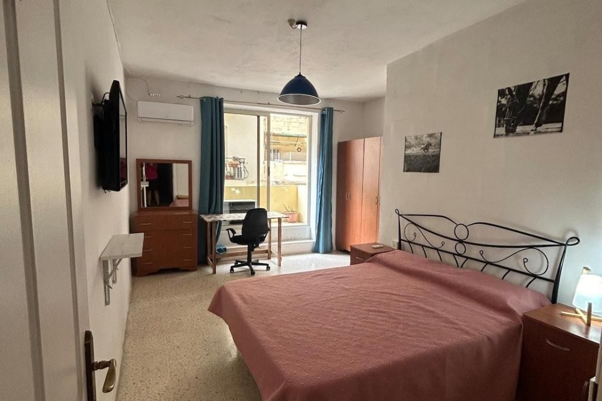 Quarto com cama, escrivaninha, cadeira, guarda-roupa e televisão na parede.