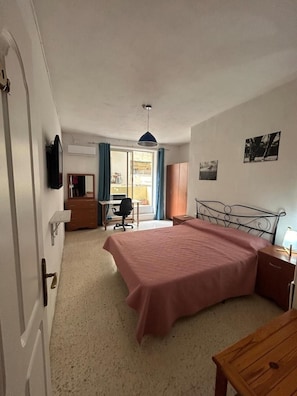 Quarto com cama, escrivaninha, cadeira, guarda-roupa e televisão na parede.