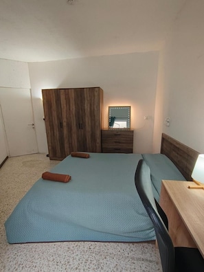 A bedroom with a bed, a wooden wardrobe, a bedside table with a lamp, and a mirror with lights.