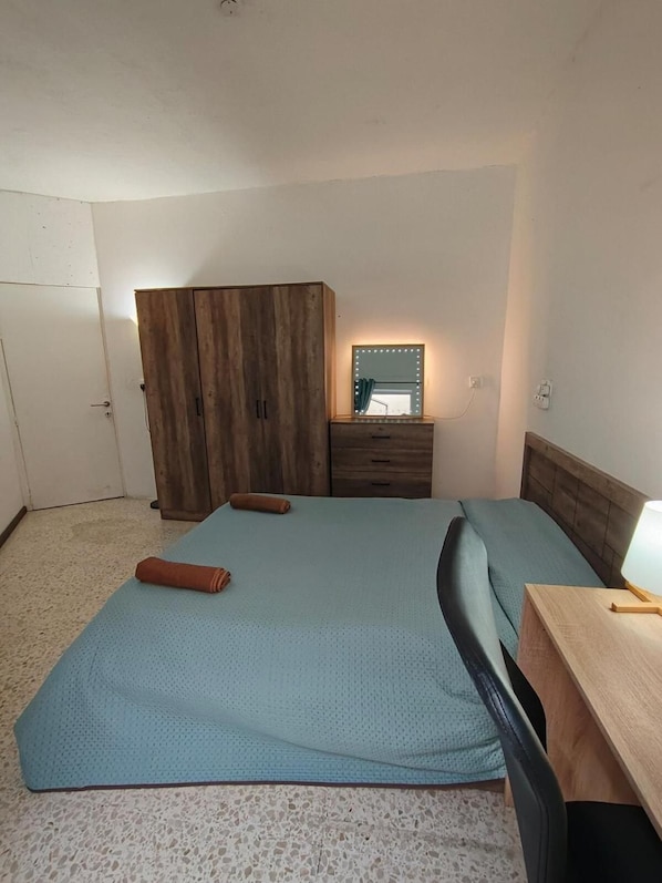 A bedroom with a bed, a wooden wardrobe, a bedside table with a lamp, and a mirror with lights.