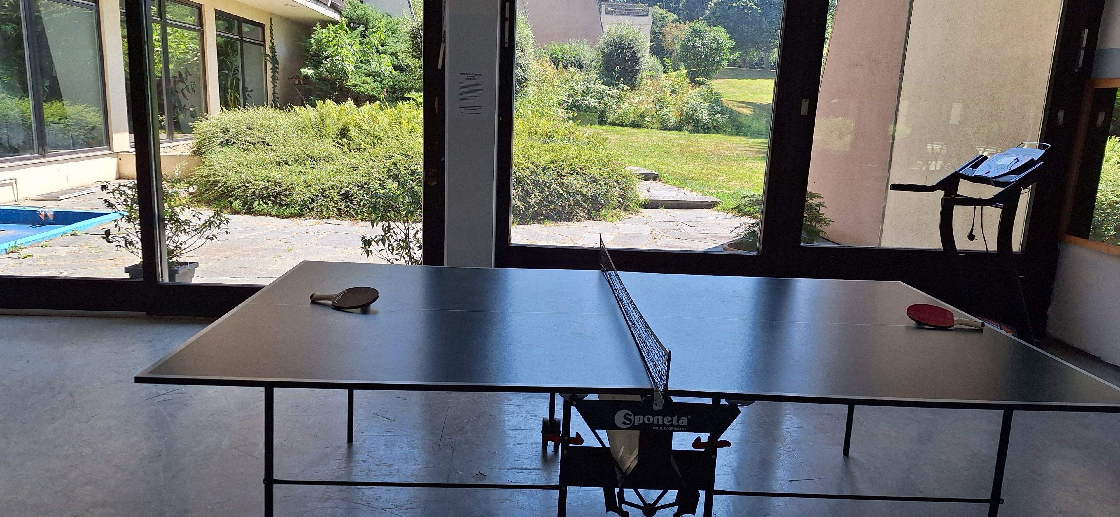 Ein Tischtennistisch mit Netz in der Mitte, ein Vogelhaus und ein kleiner Pool vor einem Gebäude mit großen Fenstern.