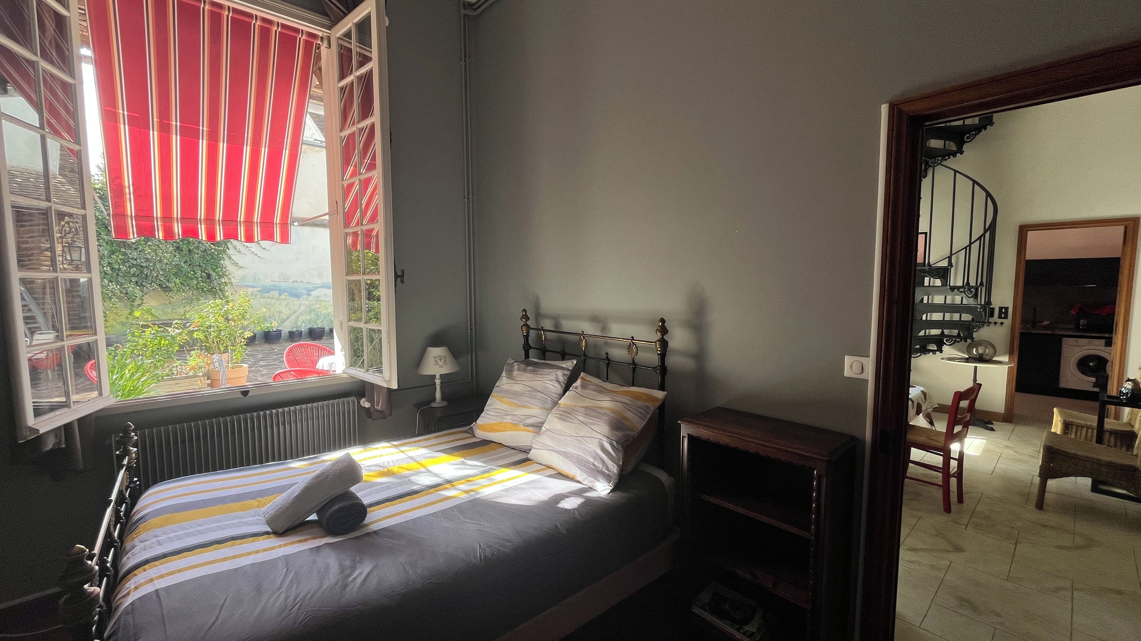 A bedroom with a bed, a window with red curtains, a radiator, and a view of a garden.