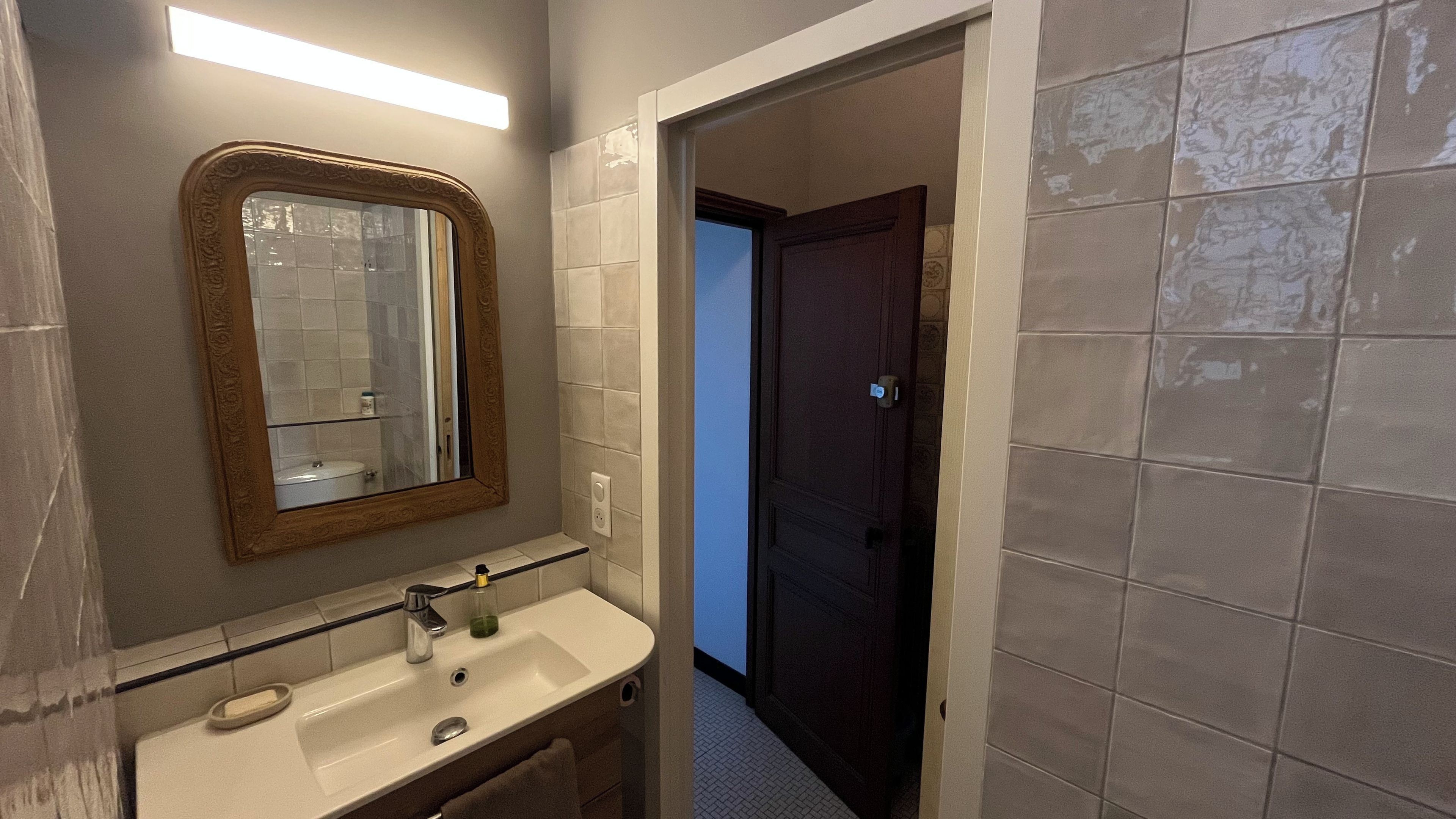 Een badkamer met een spiegel, wastafel en een deur naar een andere kamer.