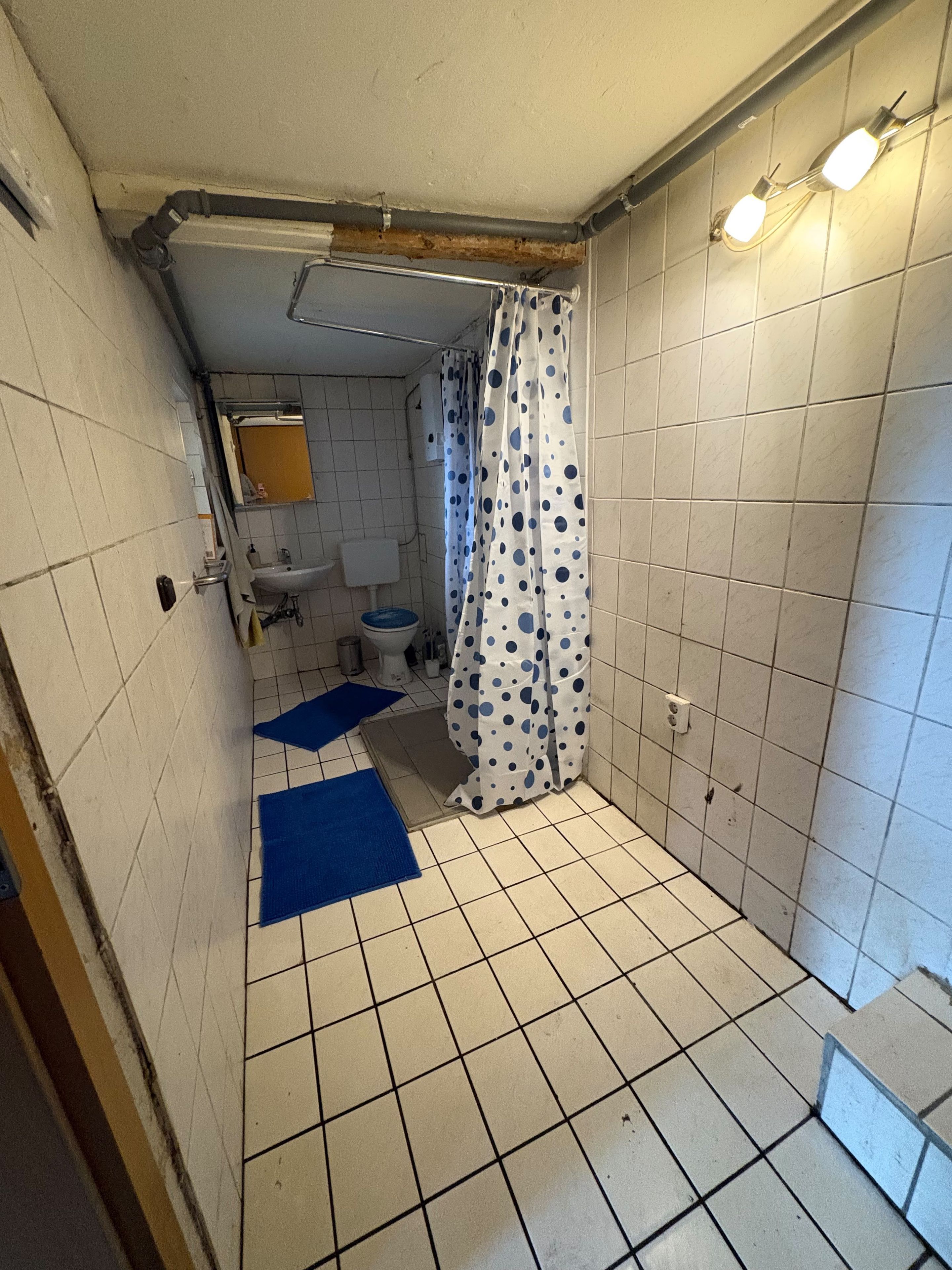 Un baño pequeño con paredes y suelo de azulejos blancos, una ducha con cortina de lunares, un lavabo e inodoro.