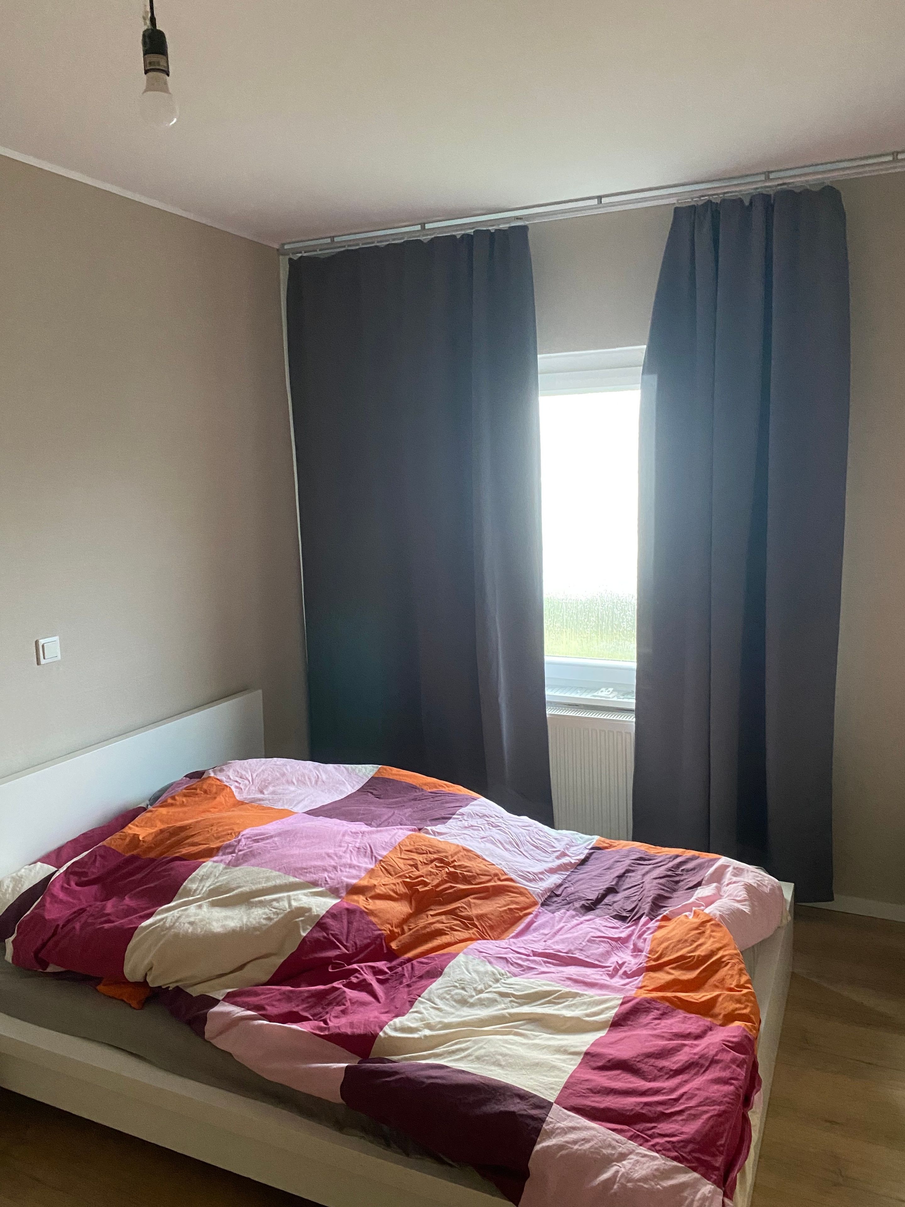 Una cama individual con un edredón colorido, una ventana con cortinas oscuras y una lámpara en el techo.
