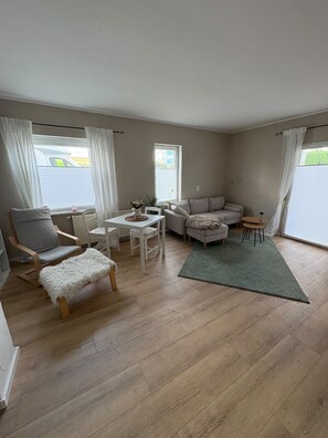 Un salón espacioso con suelo de madera, un sofá blanco, un sillón gris, una mesita redonda y una alfombra verde.