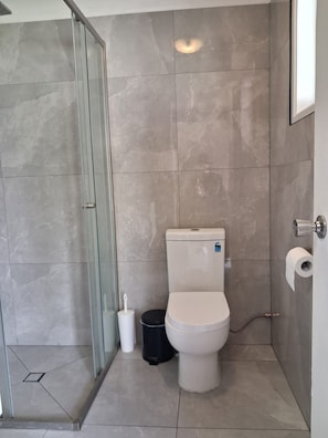 A bathroom with a toilet, a glass shower enclosure, and a black trash bin. - Centenary Motor Inn (Darra)