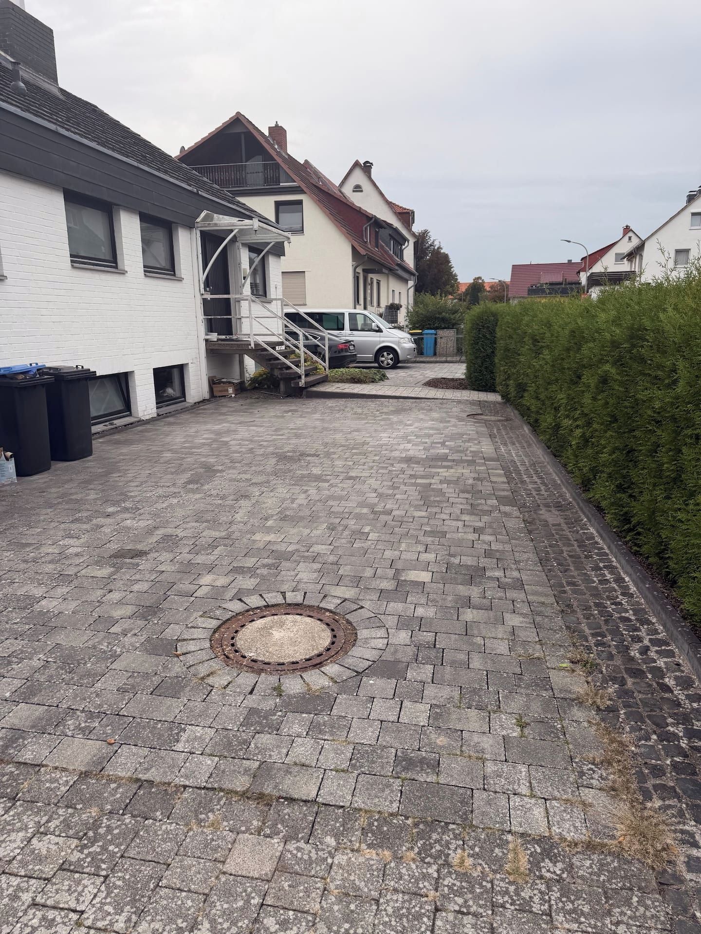 Une allée pavée mène à un quartier résidentiel où se trouvent des maisons, une voiture garée et un regard d’égout.