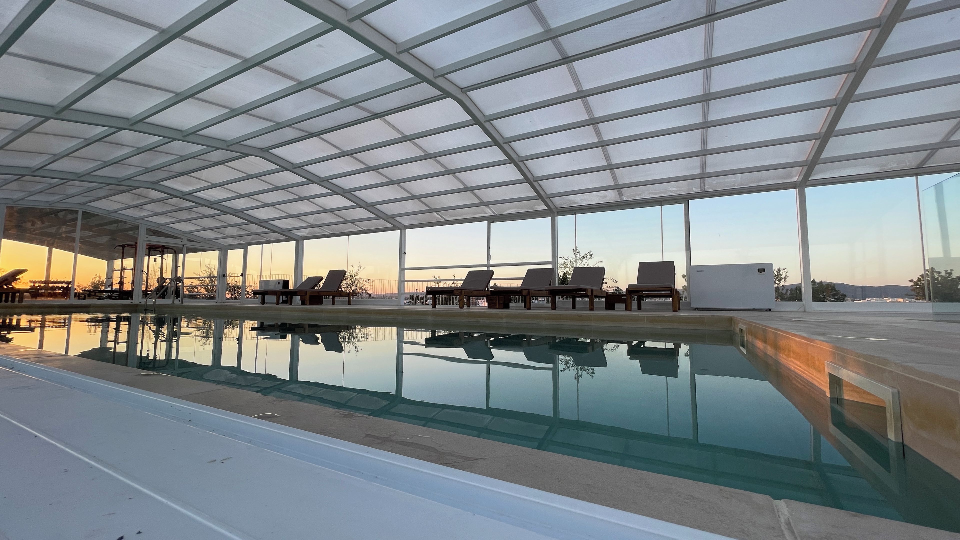 An indoor swimming pool with clear glass walls, multiple lounge chairs, and exercise equipment.