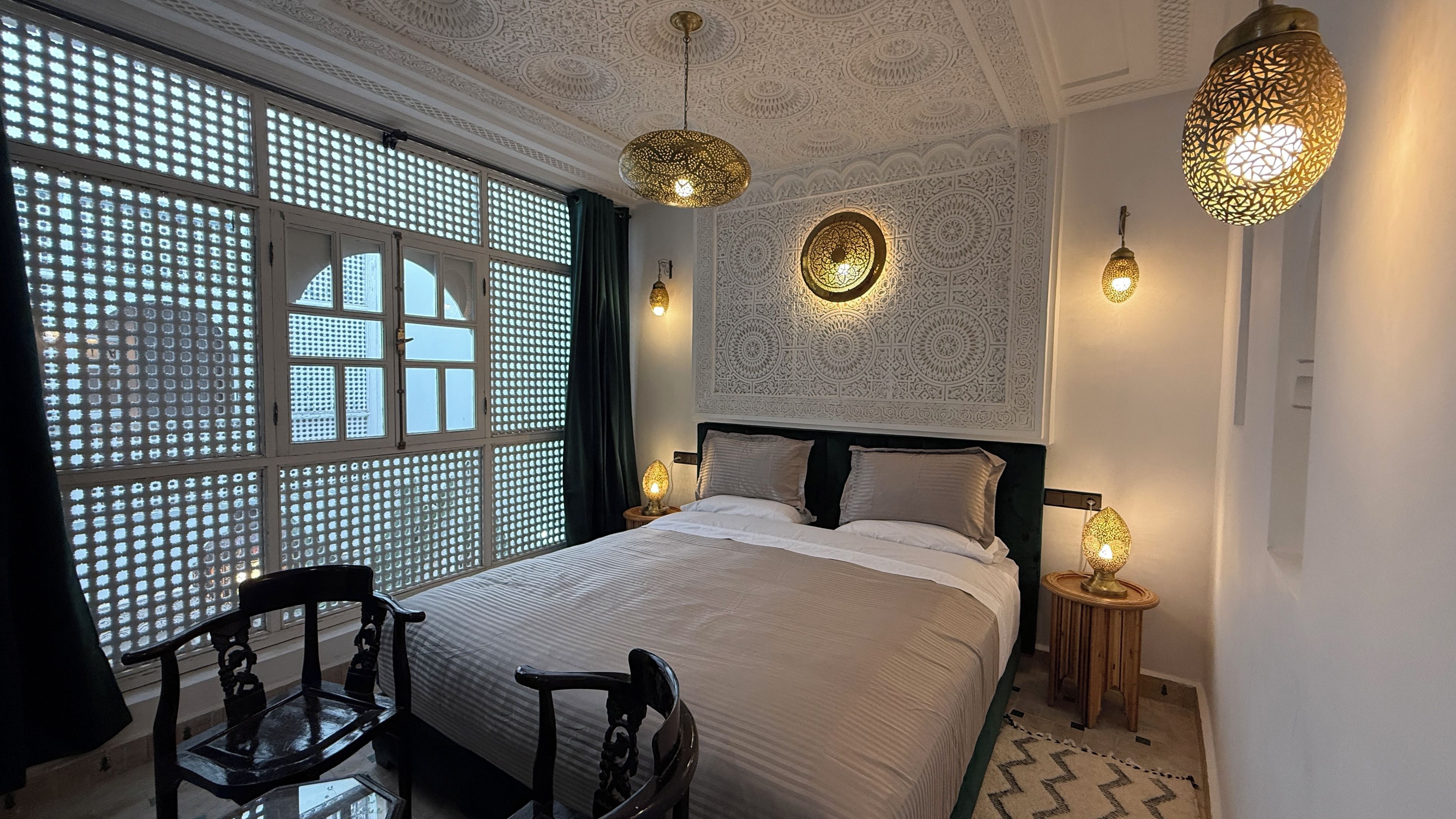 A bedroom with a large bed, a decorative wall, a window with a lattice design, and two wall-mounted lamps.
