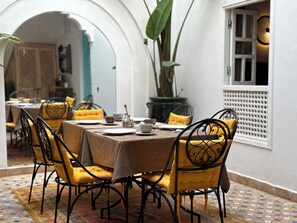 A dining area with a table set for four, surrounded by chairs with yellow cushions, a potted plant, and a view of a room with a window. - Dar 82, votre Riad privé dans la Kasbah, 4 chambres et petit-déjeuner inclus (Marrakesh)