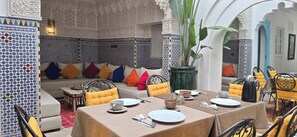 A dining area with a table set for two, a sofa with colorful cushions, and a decorative wall with intricate patterns. - Dar 82, votre Riad privé dans la Kasbah, 4 chambres et petit-déjeuner inclus (Marrakesh)