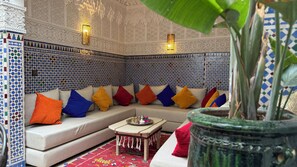A room with a patterned wall, a sofa with colorful cushions, a small table with a tray, and a large potted plant. - Dar 82, votre Riad privé dans la Kasbah, 4 chambres et petit-déjeuner inclus (Marrakesh)