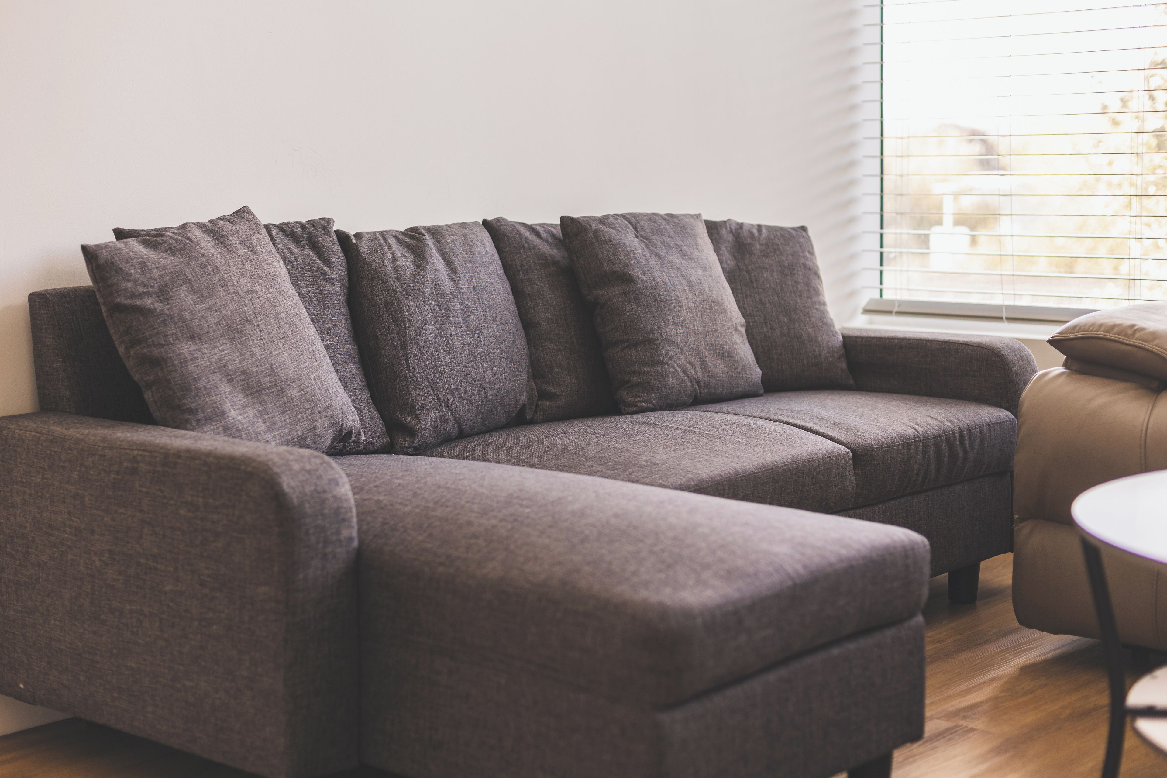 A grey sofa with multiple cushions in a room with a window and a small round table.