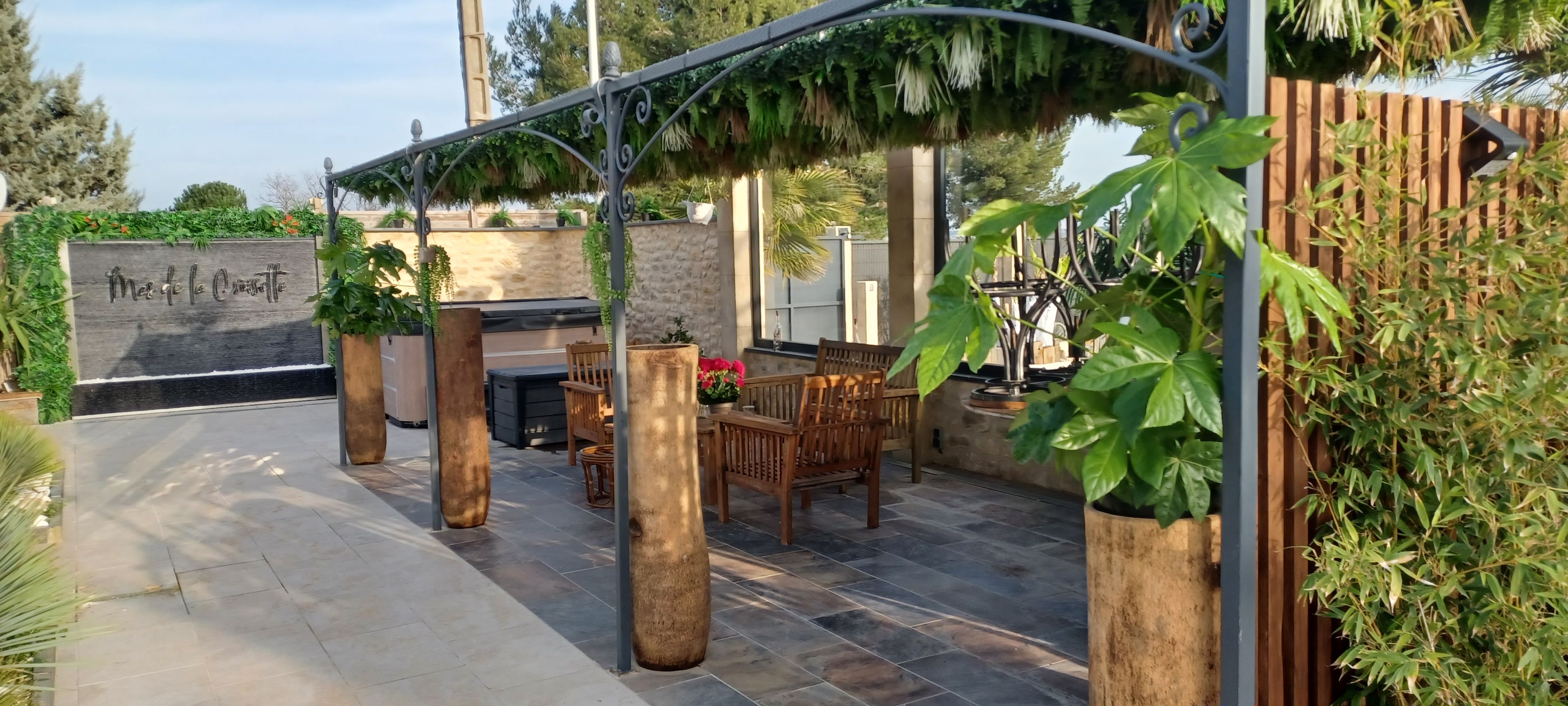An outdoor patio with a stone floor, wooden furniture, and a pergola with hanging plants.