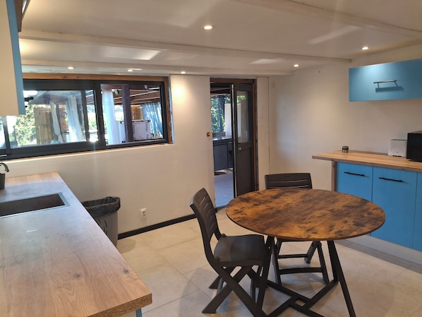 Un coin repas avec une table et des chaises en bois, une fenêtre offrant une vue agréable et un espace cuisine doté de meubles bleus et d’un four à micro-ondes.