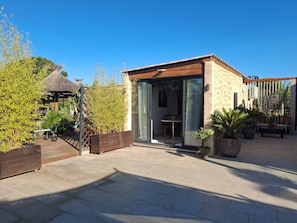Un espace patio extérieur moderne, doté d’une terrasse en bois, d’une entrée avec porte vitrée et d’un aménagement pour s’asseoir.