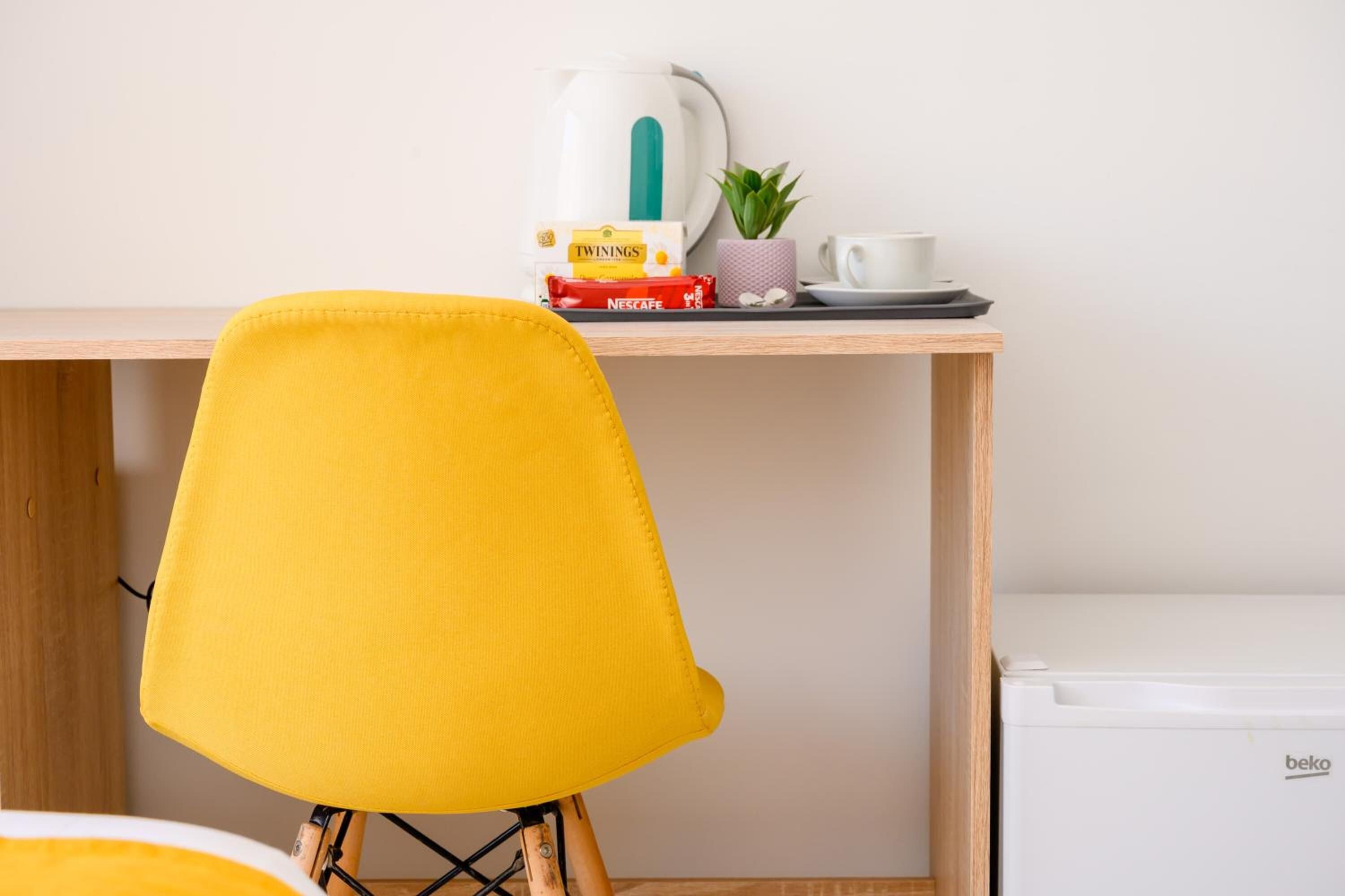 Une chaise jaune est placée devant un bureau en bois sur lequel se trouvent une bouilloire blanche, un paquet de café Nescafé, une petite plante en pot et une tasse à café blanche.