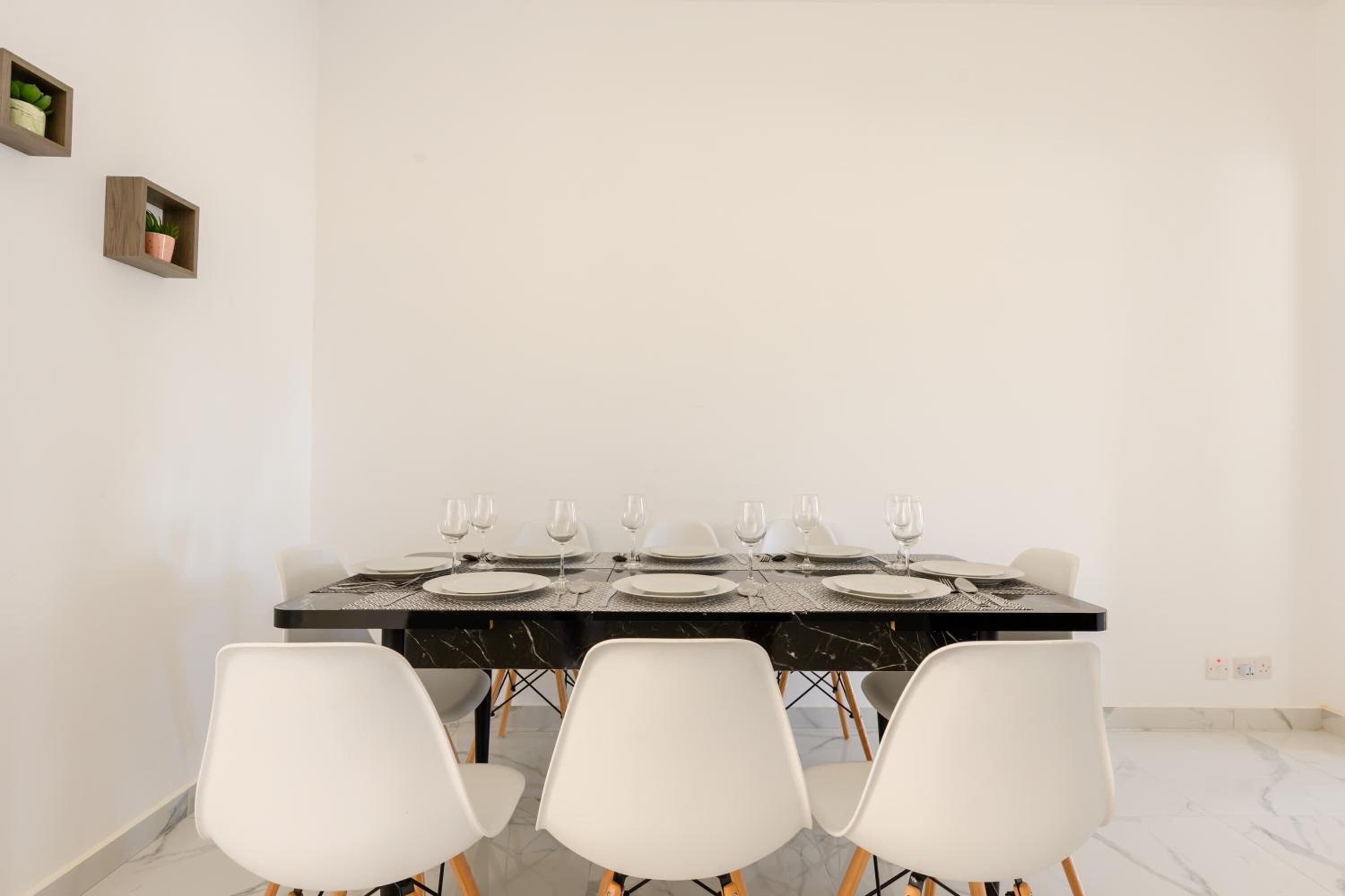 Un coin repas avec une table noire dressée pour six personnes, des chaises blanches et deux petites étagères en bois fixées au mur.