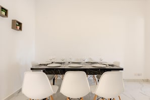 A dining area with a black table set for six, white chairs, and two small wooden shelves on the wall.