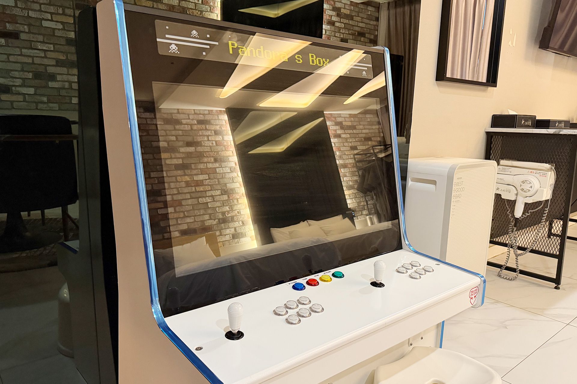 Ein Spielgerät mit Bildschirm, Joystick und Knöpfen, platziert in einem Raum mit Backsteinwand und weiß gefliestem Boden.