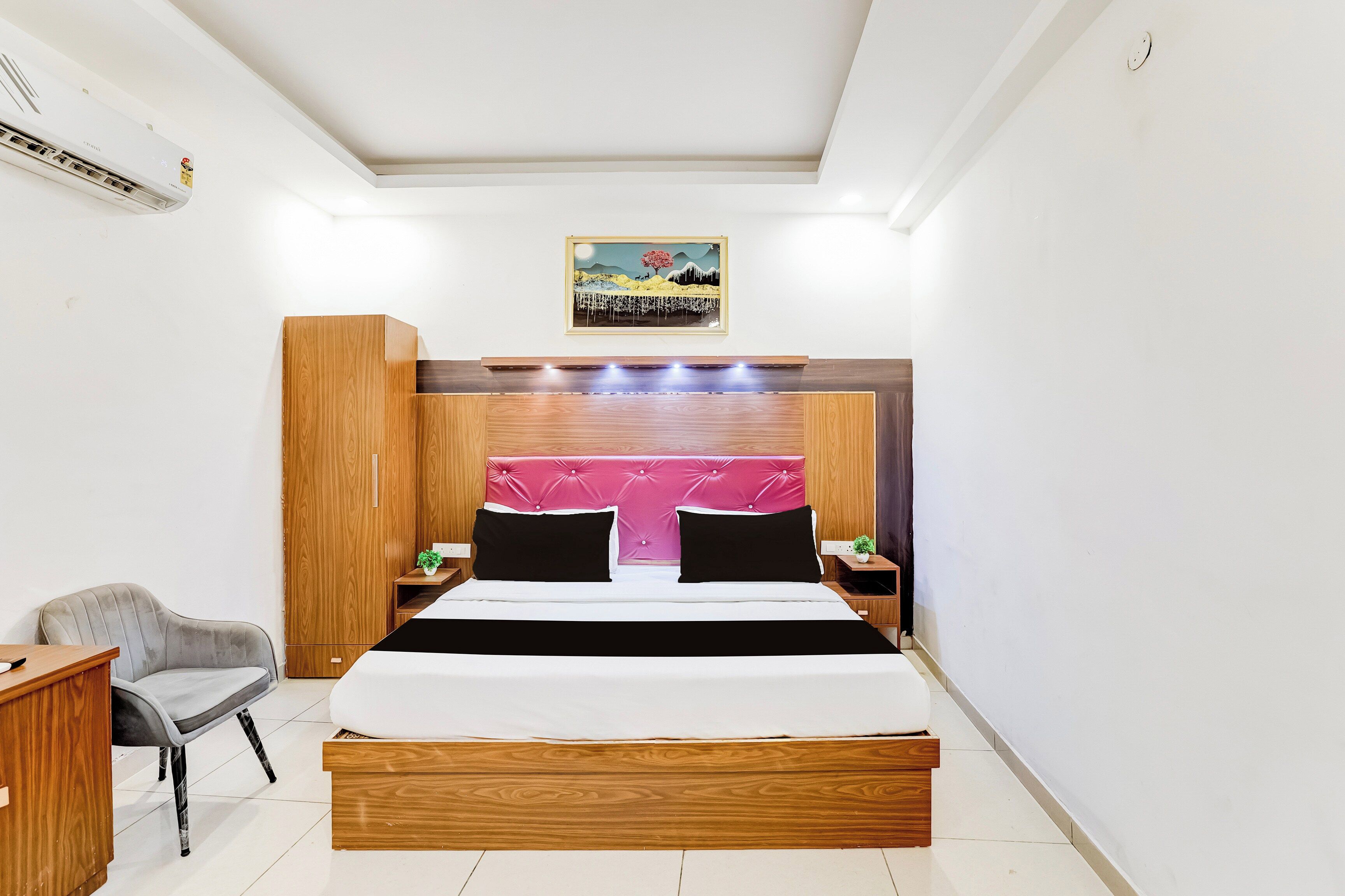 A hotel room with a bed, a chair, a nightstand, a wall-mounted air conditioner, and a painting.