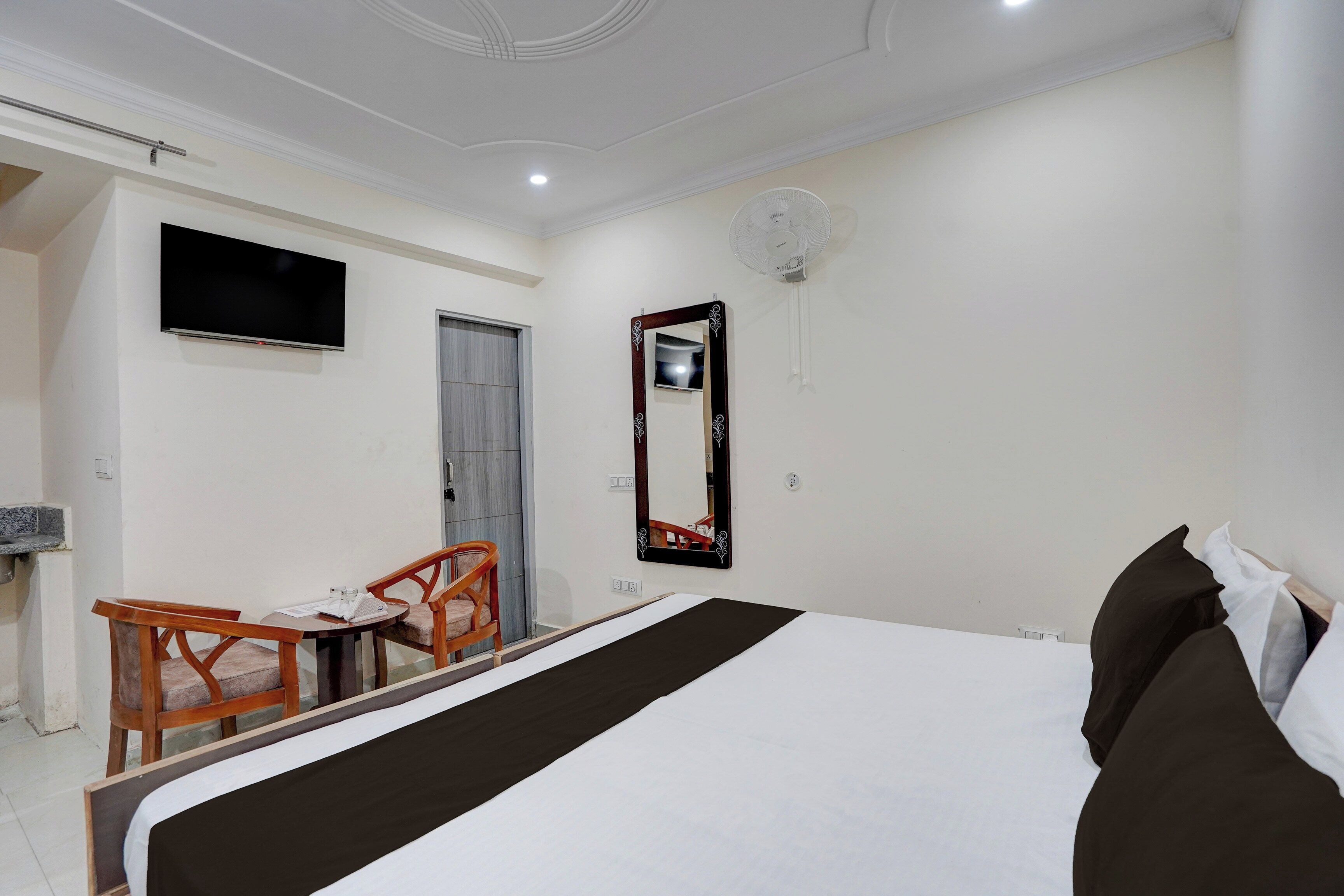 A hotel room with a bed, a desk with a chair, a TV, a mirror, and a ceiling fan.