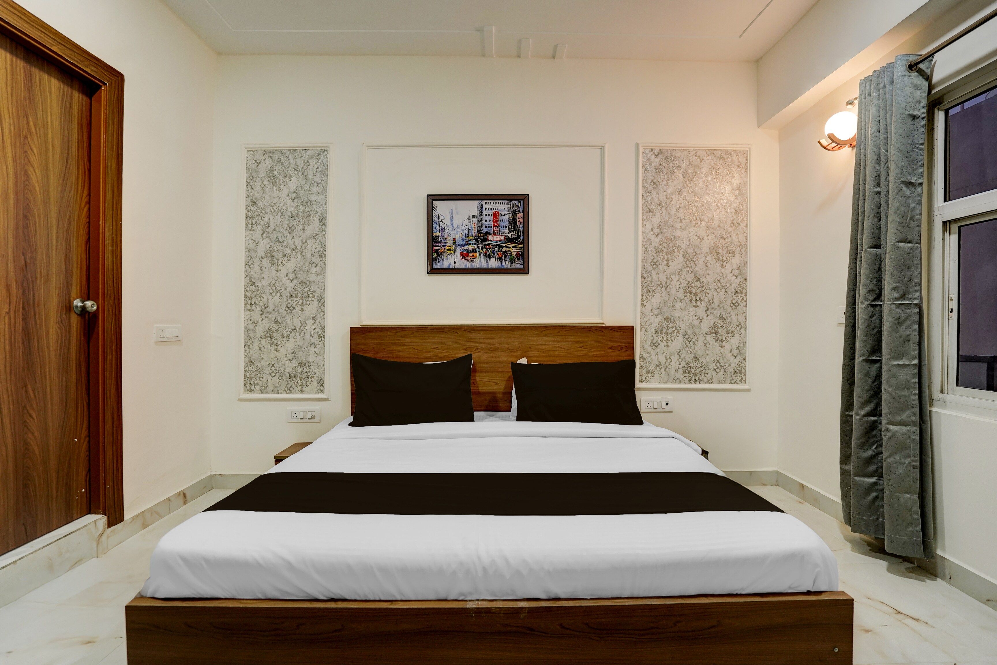 A hotel room with a wooden bed, white bedding, a framed cityscape picture on the wall, and a window with curtains.