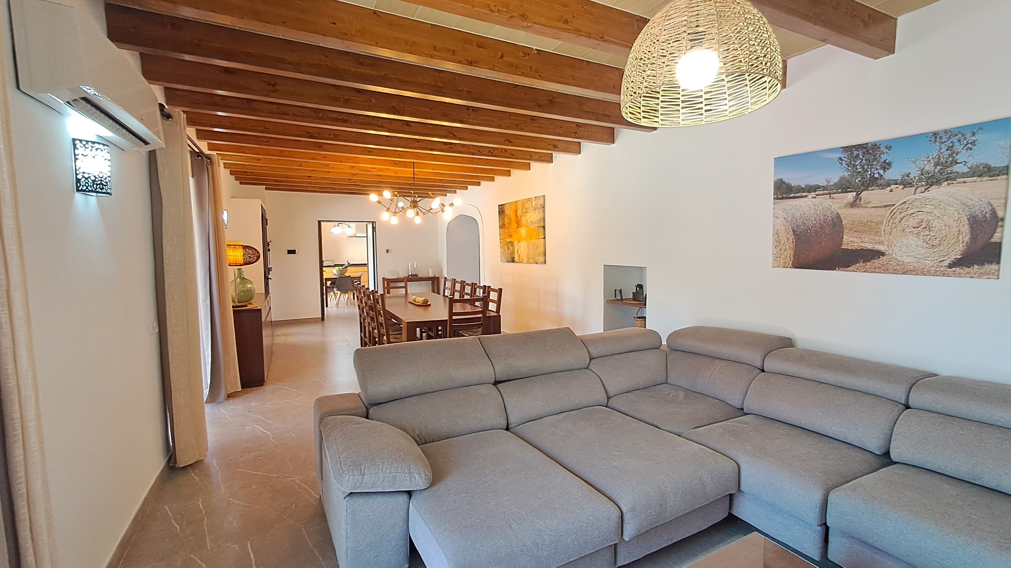 Un salon moderne avec un canapé d’angle, des poutres en bois au plafond et un coin repas à l’arrière-plan.