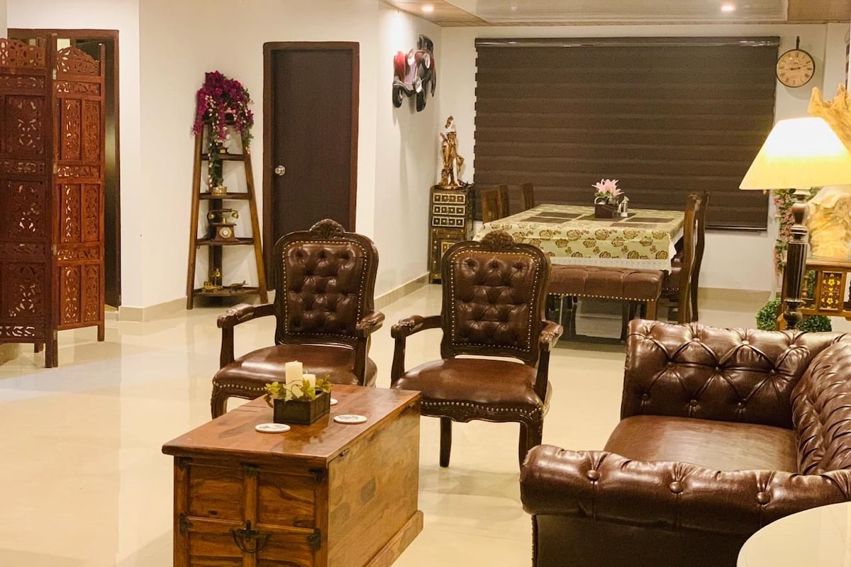 Una sala de estar con una mesa de centro de madera, sillones de cuero, un sofá, una mesa de comedor con mantel floral y un reloj en la pared.