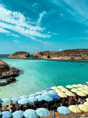 A beach with clear turquoise water, rocky formations, and umbrellas. - RODOLFU F5A (Tas-Sliema)