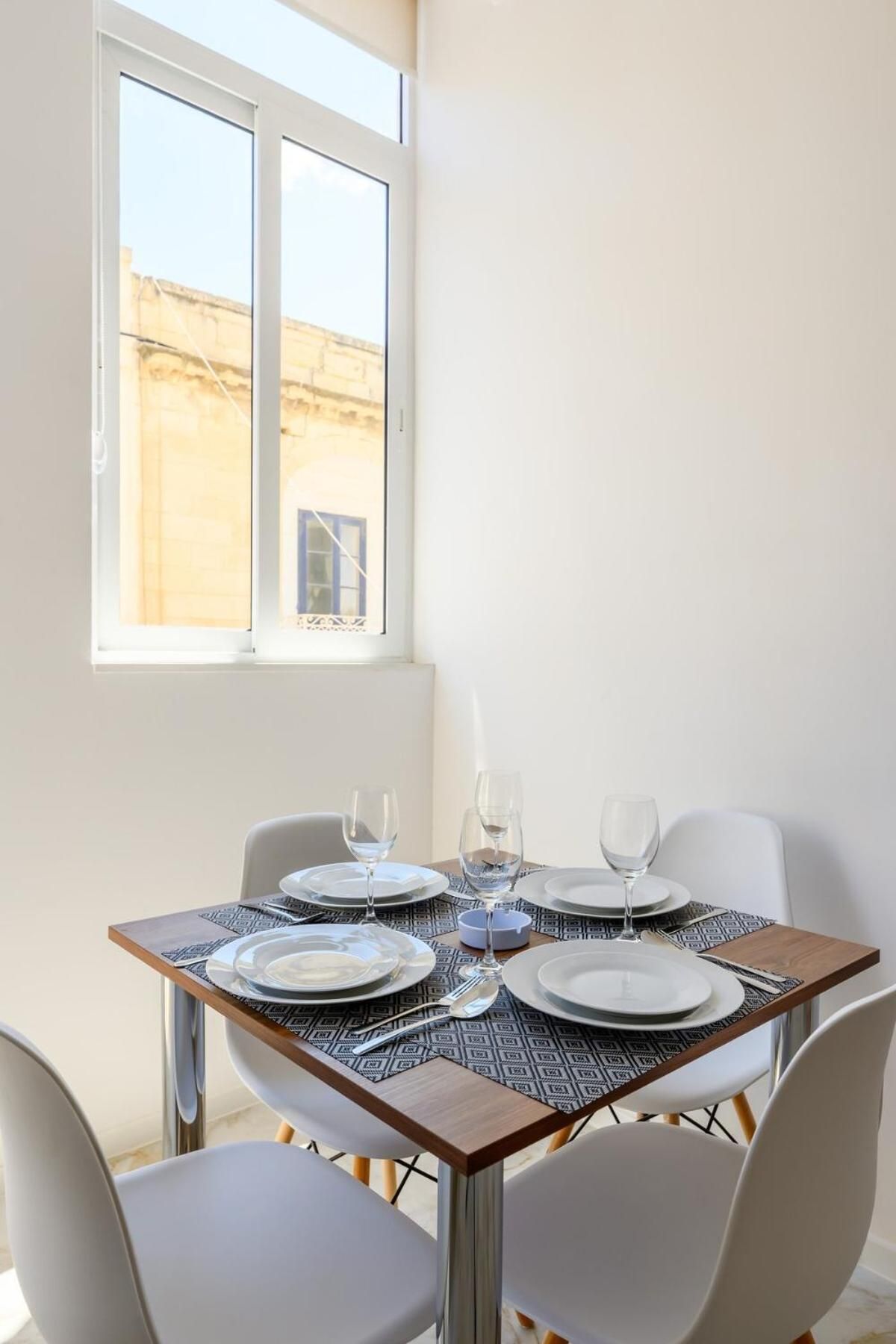 Mesa para dos, dispuesta con platos, vasos y cubiertos, cerca de una ventana con vistas a un edificio.