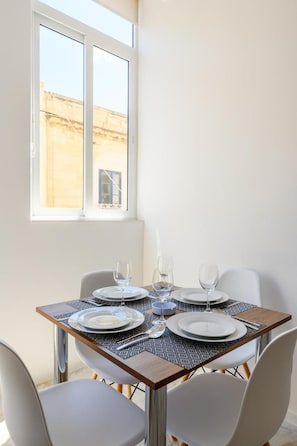 A dining table set for two with plates, glasses, and cutlery, positioned near a window with a view of a building. - RODOLFU F5A (Tas-Sliema)