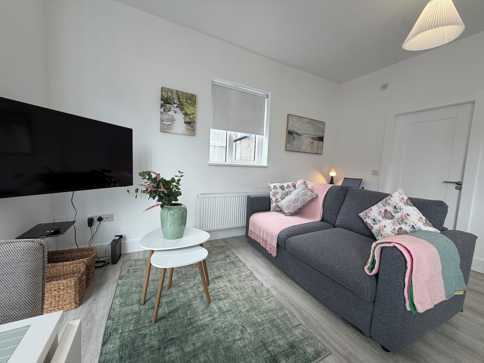 A modern living room with a flat-screen TV, a grey sofa with decorative pillows, a small white table with a green vase, and a window with a view of a stream.