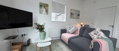 A modern living room with a flat-screen TV, a grey sofa with decorative pillows, a small white table with a green vase, and a window with a view of a stream.