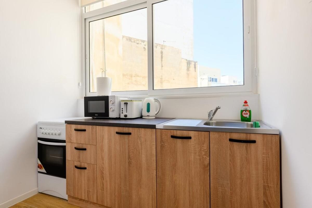 Cocina compacta con microondas, hervidor y una botella de detergente para platos sobre la encimera.