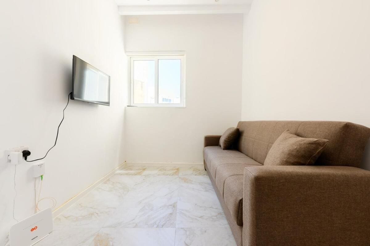 Quarto com sofá, televisão de tela plana na parede, janela e tomada com aparelho conectado.